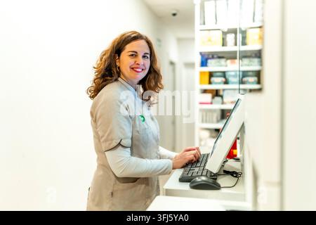 Smiling woman pharmacist supporting customers and managing inventory using a computer system in a modern drugstore, ensuring efficient healthcare serv Stock Photo