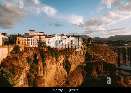 Whitewashed buildings of Ronda's historic center perched precariously on a towering cliff edge, illuminated by warm golden hour light under a vast sky Stock Photo