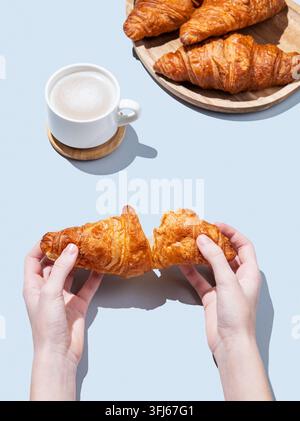 Hands breaking a fresh croissant over a blue background with a cup of cappuccino or latte coffee and pastry. Creative mockup for cafe advertising or F Stock Photo