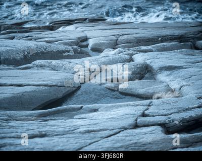A rocky shoreline displays unique textures and shapes created by water erosion. Pools of water are visible among the rocks during low tide. Stock Photo
