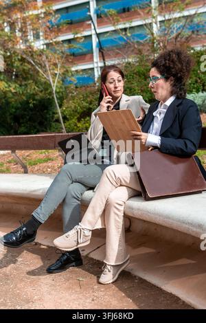 Two professional businesswomen collaborating and discussing work topics during an outdoor meeting, using a smartphone and tablet for communication and Stock Photo