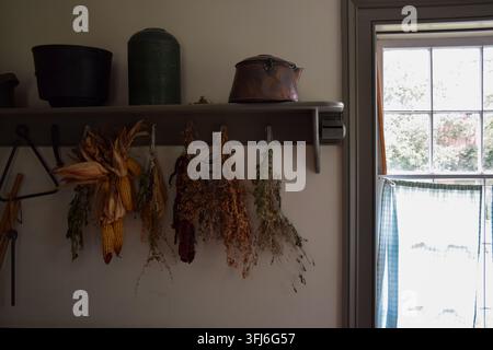 Dried herbs and corn cobs hanging from hooks under shelf with vintage metal canisters and window with checkered curtain in rustic country kitchen Stock Photo