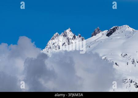 The photo shows snow-capped mountain peaks against a cloudless bright blue sky. Mountains occupy a significant part of the frame. The peaks are covered with a thick layer of snow, which has a heterogeneous structure: both smooth, sunlit areas and dark-colo Stock Photo