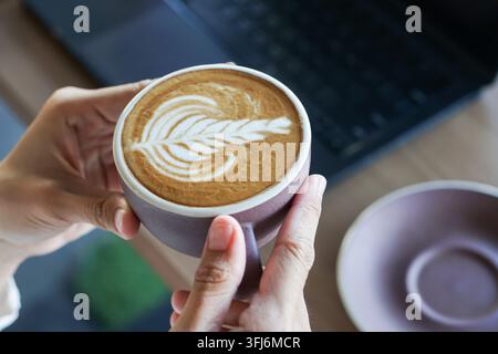 Close-up of hands holding a cup of latte with leaf foam art beside a laptop and empty saucer, creating a cozy work break scene. Stock Photo