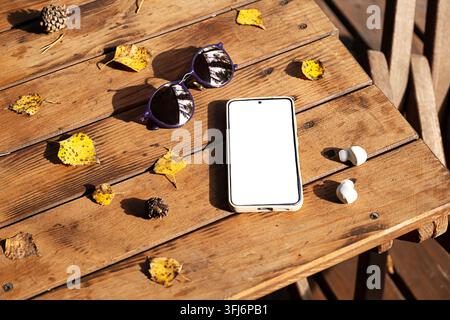 Smartphone with blank screen, sunglasses and wireless earbuds on wooden table with yellow autumn leaves. Fall flat lay composition in sunlight. Stock Photo