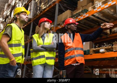 A diverse warehouse team collaborates on logistics, wearing safety gear. They focus on efficiency and teamwork amidst industrial shelving units. Stock Photo