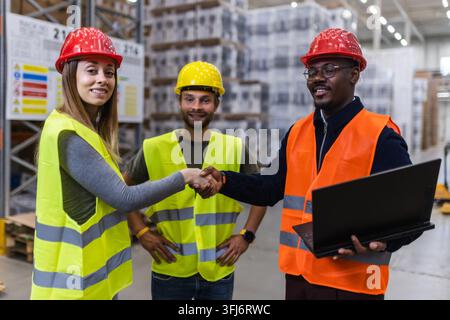 A group of engineers in a warehouse collaborating on logistics. They are wearing safety gear and shaking hands, demonstrating teamwork and coordinatio Stock Photo