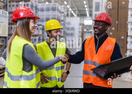 Three warehouse workers wearing safety gear discuss logistics in an industrial setting. They appear to be sharing ideas and collaborating on a project Stock Photo