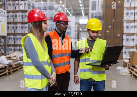 Three warehouse workers in reflective vests and helmets discuss logistics using a laptop. They are analyzing operations and ensuring efficiency in a b Stock Photo