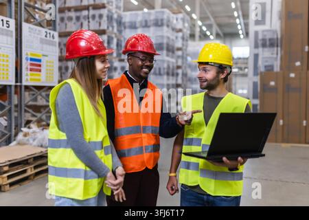A group of workers in safety gear collaborates in a warehouse, using a laptop to review logistics data, showcasing teamwork and technology in an indus Stock Photo