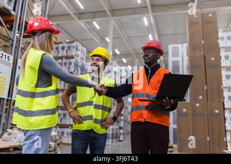 Three warehouse workers in safety gear are standing in an aisle, shaking hands, and using a laptop to discuss operations. Stock Photo