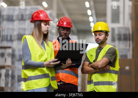 Three warehouse workers in safety gear discuss project details using a laptop in a large storage facility. The scene emphasizes teamwork and industria Stock Photo