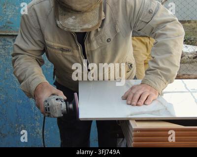 A man is grinding the edge of a large-format tile using an angle grinder with a special attachment. The tiler is working outdoors, carefully steadying Stock Photo