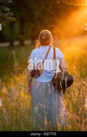 Back view of middle aged female photographer standing in meadow at sunset with camera bag over her shoulder, looking toward setting sun in warm golden Stock Photo