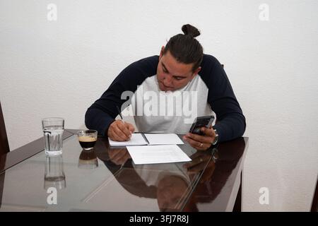 Young man telecommuting at home, writing notes and checking his phone at a glass table Stock Photo