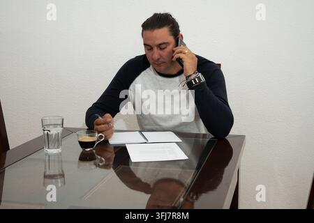 Man talking on mobile phone while writing notes at a home office table Stock Photo