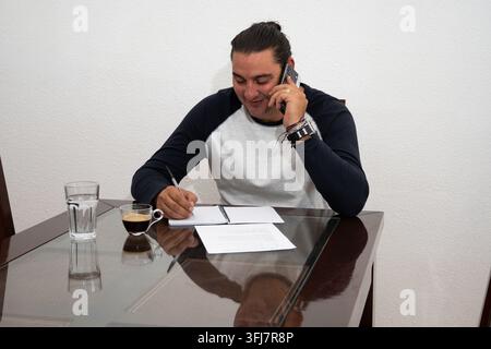 Man working from home, talking on phone and writing notes at table Stock Photo