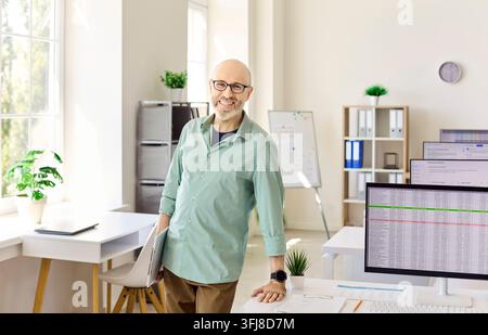 Smiling middle aged teacher in modern computer classroom, standing with laptop Stock Photo