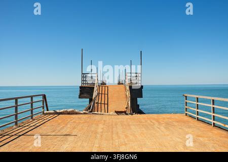 Old concrete pier with harsh shadows from rusted metal railings extends into blue calm sea water against clear sky on bright sunny day. Stock Photo