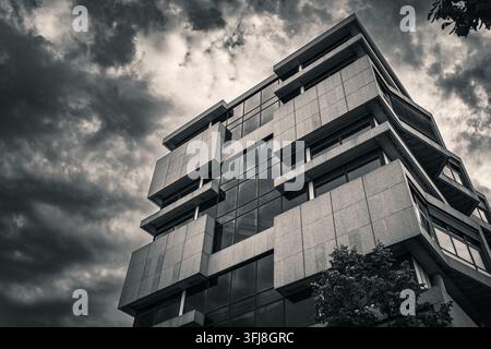 Black and white low angle view of a geometric concrete office building beneath dark dramatic clouds. Strong architectural composition suitable for bus Stock Photo
