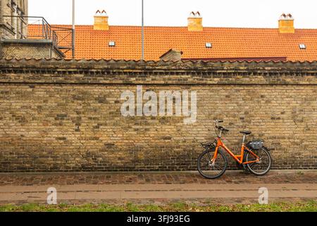 Copenhagen, Denmark - December 16, 2023: Orange bicycle standing on a sidewalk next to a textured brick wall under a tiled roof Stock Photo