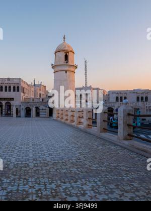 Historic round stone minaret standing beyond patterned tiled plaza with traditional plaster fence under clear sky in Souq Waqif. Stock Photo