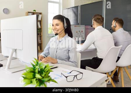 Smiling female call center operator with headset working in office with team Stock Photo