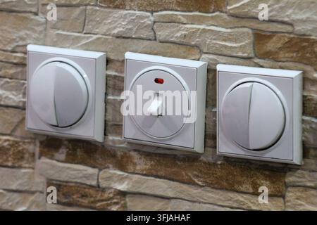 Close-up of three white electrical light switches with a red indicator light, mounted on a textured beige stone wall. Stock Photo
