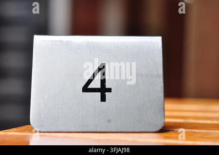 A silver brushed metal table number stand displaying the numeral four. It is positioned on a wooden surface with a blurred dark and brown background. Stock Photo