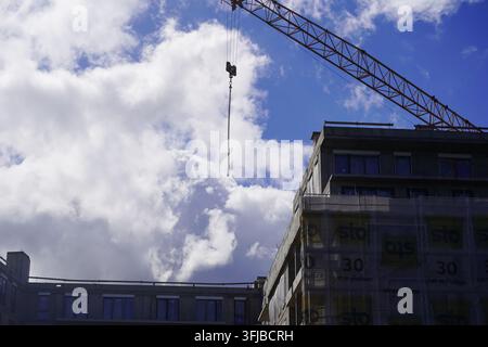 Tall construction crane operating above a modern building under development against a bright blue sky filled with white clouds. Stock Photo
