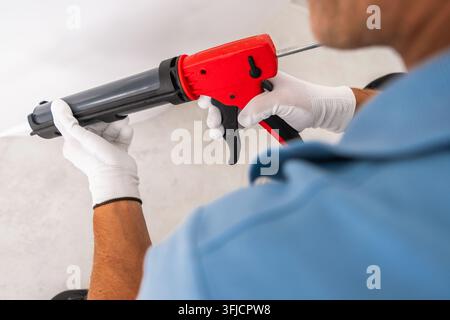 Close view of a worker holding a cartridge caulking gun with both gloved hands during bathroom renovation. The applicator and trigger are the main foc Stock Photo