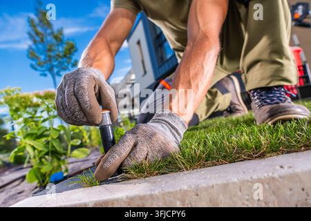 Close view of a gardener adjusting the head of a pop up sprinkler at the edge of a lawn. Gloved hands hold the irrigation fixture beside the garden bo Stock Photo