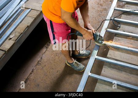 A man using electric grinder to cut metal frame in workshop with sparks and protective footwear Stock Photo