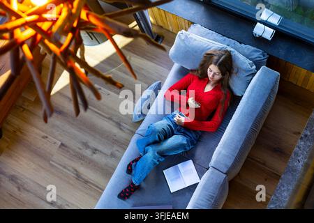 Young Woman Relaxing on Sofa and Using Smartphone. Young woman in a red sweater reclines on a gray sofa using her smartphone, with an open book beside Stock Photo
