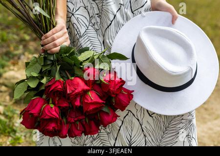 Person Holding Red Roses and White Hat Outdoors. Person in a patterned dress holds a large bouquet of red roses and a white hat with a black band Stock Photo