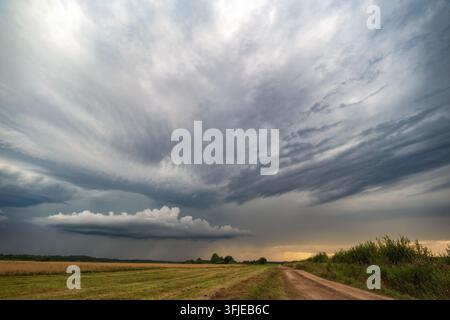 Dramatic storm clouds spreading across the sky above agricultural fields, with a rural dirt road leading toward the distant horizon at sunset Stock Photo