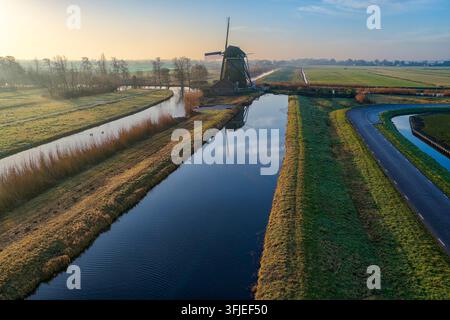 Rietveldse windmill standing proud in the Dutch polder landscape early in the morning, with light reflecting off the calm canal water and green fields in Hazerswoude-Dorp Stock Photo