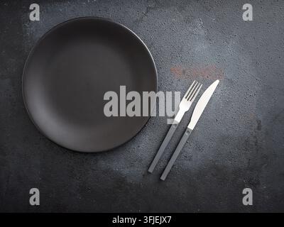 Empty black plate and modern cutlery resting on a dark grunge table. Preparing a minimalist dining setting with copy space Stock Photo