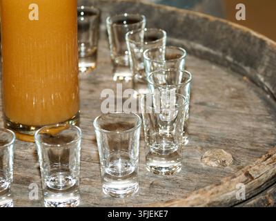 Empty and partially filled shot glasses with a tall glass of orange liquid on a rustic wooden barrel. Stock Photo