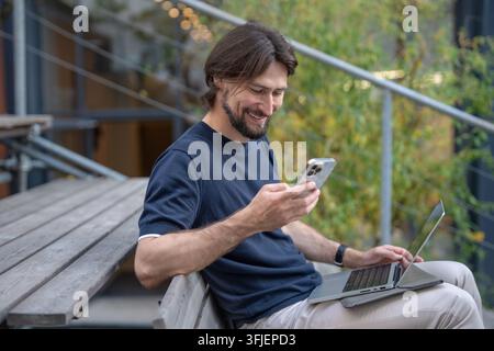 Smiling businessman using smartphone and laptop while working remotely outdoors in urban setting. Stock Photo