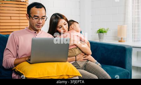 In their home office a mother carries her sleeping daughter while her husband works on a laptop showcasing the art of balancing family work and respon Stock Photo