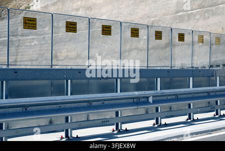 Steel guardrail and wire mesh fence protecting a road, displaying multiple yellow warning signs indicating danger from high voltage electricity, provi Stock Photo
