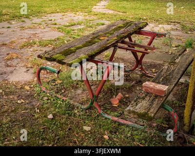 Old broken and decayed wooden picnic table with moss and rust in an abandoned park area overgrown with weeds Stock Photo