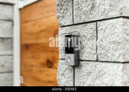 Close up of a smart intercom video doorbell camera mounted on a grey stone brick wall. Modern private house entrance door with wood panel background. Stock Photo