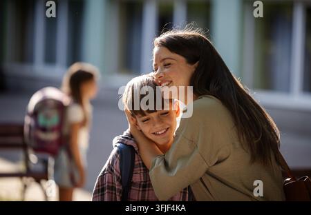 Mother and son hug outside a school during the afternoon. The older children standing in background in courtyard. Stock Photo