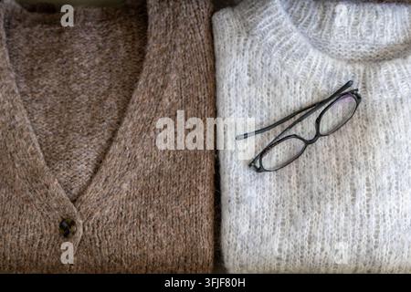 Folded cardigan and sweater display side by side with eyeglasses resting on the fabric. Concept of cozy autumn wardrobe styling and knitwear presentat Stock Photo