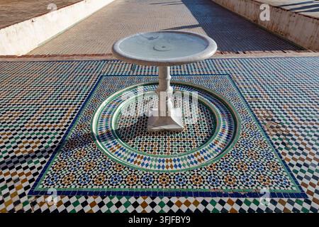 A stone fountain centered on an intricate mosaic floor in Marrakesh, Morocco, surrounded by geometric patterns under bright daylight. Stock Photo