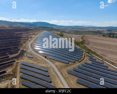 Solar panels line rural hillside rows, forming large power array beside dirt roads. Bright sunlight reflects from modules, and renewable energy site s Stock Photo