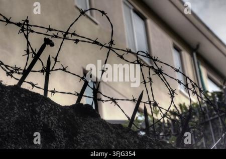 Close-up of a brick wall topped with sharp barbed wire and metal spikes under a cloudy sky, symbolizing security, restriction, and protection. Stock Photo