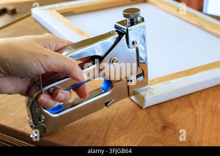 Staple gun in hand with a chrome tacker fixing canvas to a wooden stretcher frame on a table, illustrating artwork stretching process. Stock Photo